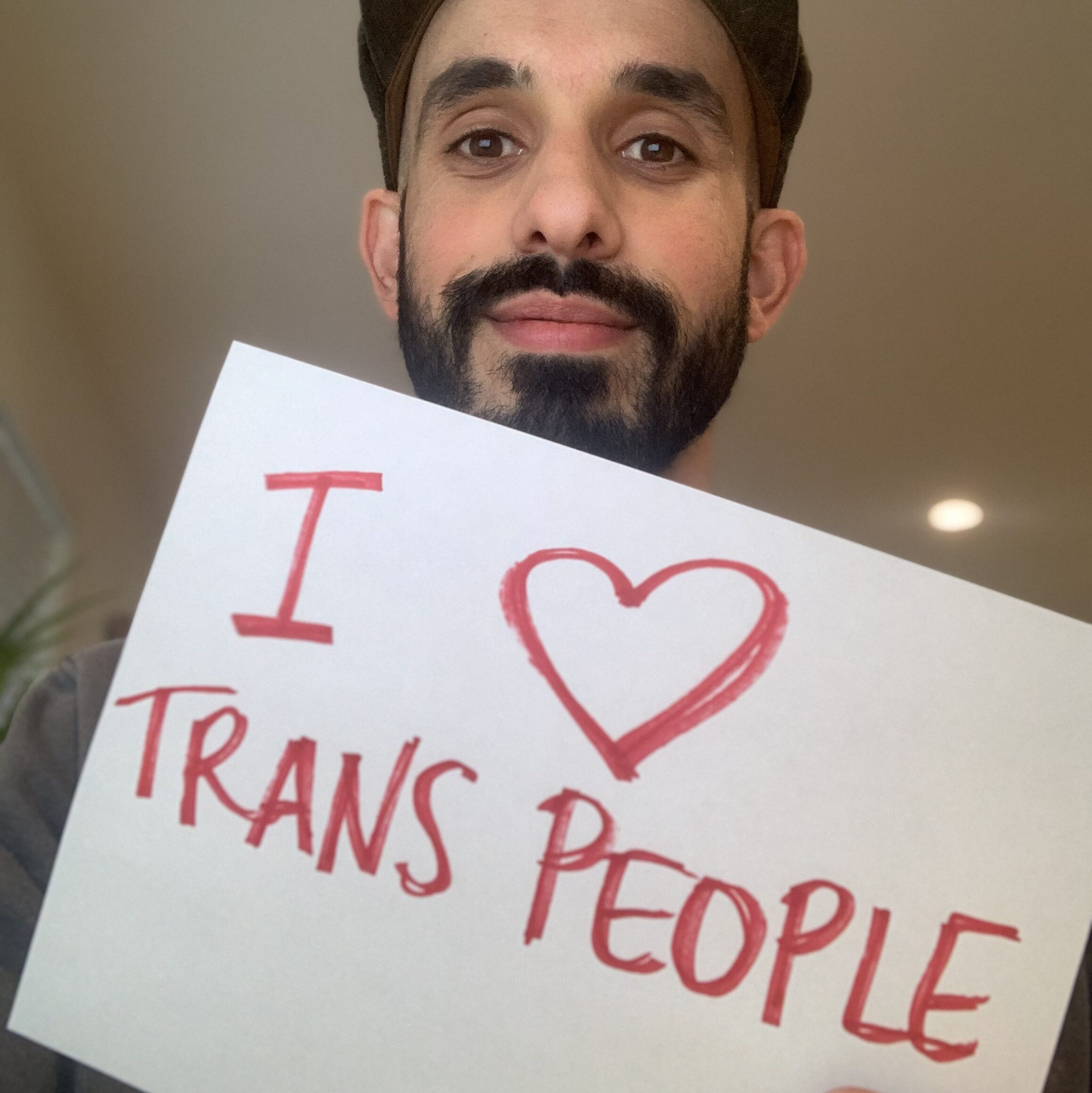 Eric Garcia, a light-skinned Latinx person with a thick black mustache and beard holds a handwritten sign that says "I (drawn heart) Trans People”