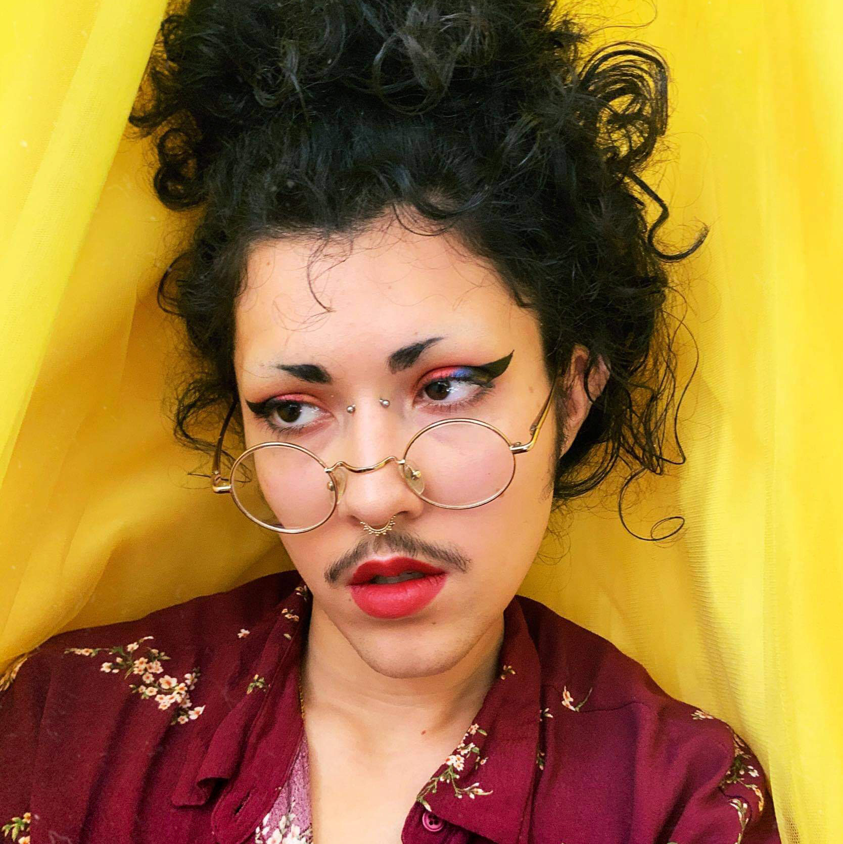 a bust view of art twink, a thin, light-skinned Bengali-American on a yellow fabric backdrop. their long curly black hair is in a messy bun on top of their head, with a clean-shaven face except a mustache & eyebrows shaved into dog brows. they are wearing winged eye-liner with red & blue eyeshadow & red lipstick. they have a bridge & septum piercing with small round & gold glasses & a purple floral unbuttoned shirt. 