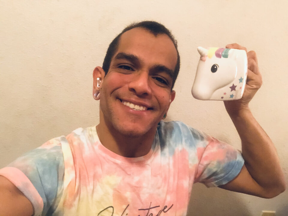 JanpiStar, Latinx Puerto Rican holding with their left hand a cup in the shape of a unicorn. JanpiStar is smiling and is wearing a tie dye shirt.