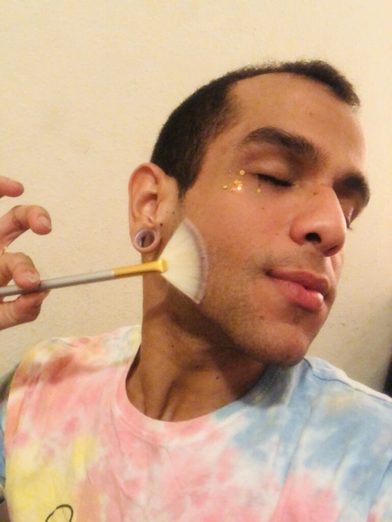 JanpiStar, light brown skin latinx holding with their fingers a makeup brush against their face. Janpistar have their eyes closed and glitter in the face. They is wearing a colorful tie dye shirt.