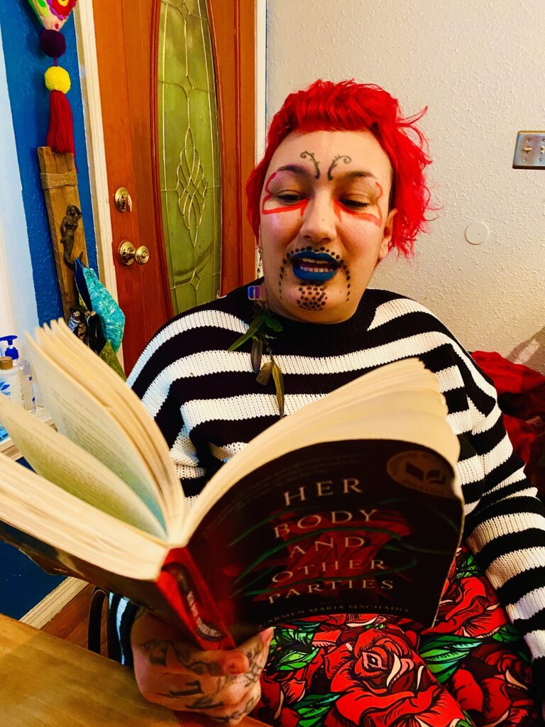 A portrait of NEVE, brown-skinned and bright red-haired, tattooed antenna at his third eye practically trembling, mid-sentence, teal painted lips pulling up to reveal cream-colored teeth, reading aloud from a book called Her Body and Other Parties by Carmen María Machado. NEVE is wearing a black and white horizontally striped sweater, leggings printed with an art deco/mosaic-like graphic of red roses and green leaves, and their face is decorated with petal-shaped lines beneath their eyes and a goatee made of dots.