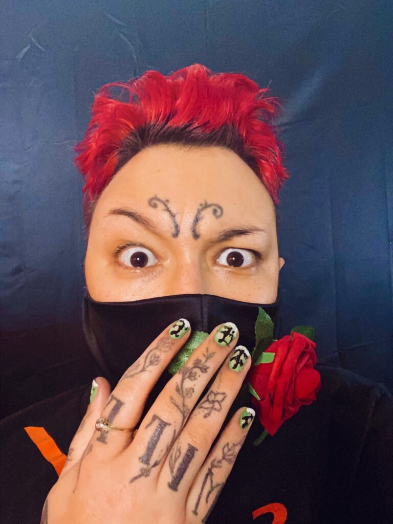 A close-up selfie of NEVE, brown-skinned, bright red hair climbing into a pomp, face tattoos centered between the rise and fall of expressive, slapstick eyebrows. He is wearing a black silk mask covering his nose and mouth onto which a three-dimensional cloth red rose is stitched. This mask is the emblem of La Casa de Montague, in Seattle Shakespeare's new, filmed, bilingual production of Romeo y Julieta, adapted by Ana María Campoy from the Shakespeare text, directed by Sophie Franco, choreographed for film by Alyza Del Pan Monley. NEVE is covering his already covered mouth as if in shock and surprise with a messily yet exquisitely lacquered hand, each fingernail reminiscent and expressive of a small snowy forest in the Pacific Northwest. https://www.seattleshakespeare.org/romeoyjulieta/